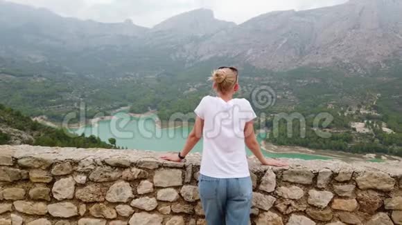 一个年轻的女人从一座古老城堡的墙上看到一个美丽的风景有一个蓝色的湖和山视频的预览图