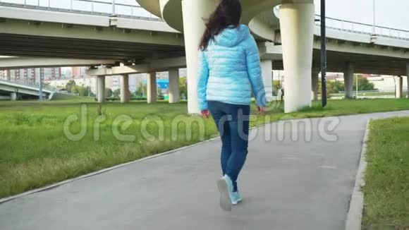 年轻女子穿着牛仔裤和蓝色夹克在现代城市的高速公路背景下行走在城市散步的后景妇女视频的预览图