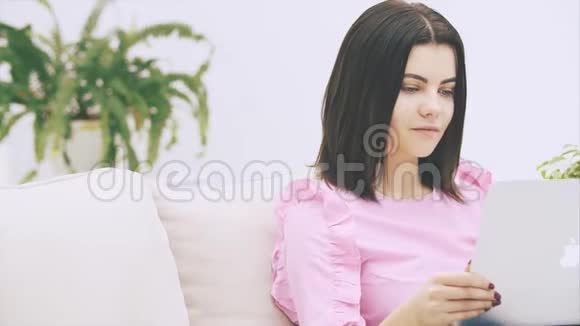可爱的白种人黑发女孩坐在沙发上拿着笔记本电脑在互联网上冲浪对情绪异常的事情做出反应视频的预览图