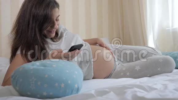 怀孕的白种人女人躺在床上使用智能手机和戳她的胃视频的预览图