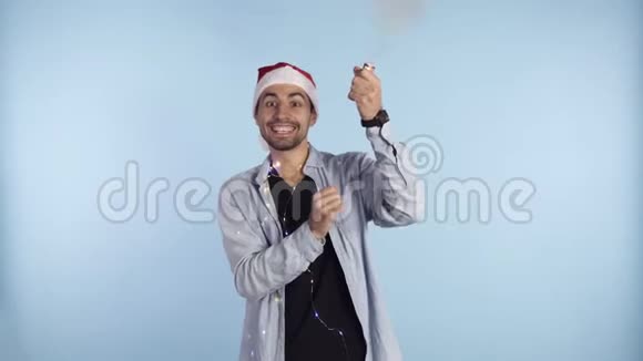 年轻的微笑的男人穿着休闲服头上戴着圣诞老人帽蓝色背景上爆炸的纸屑饼干视频的预览图