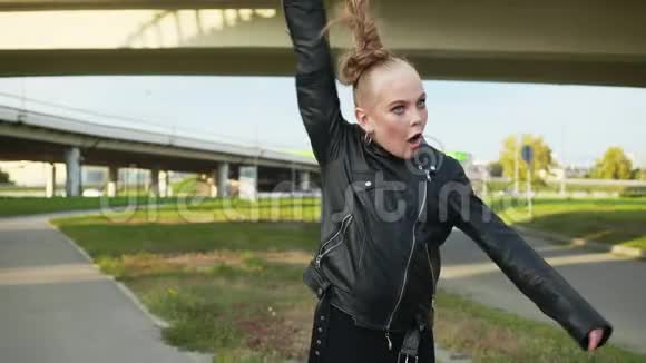 年轻女孩穿着黑色皮夹克在城市景观背景上抱着发尾戴着前摄像头的摇滚女孩视频的预览图