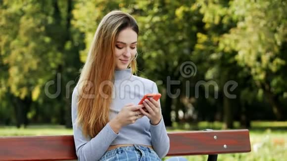 一张可爱的红头发年轻女子在公园的长凳上用手机特写的肖像年轻女子微笑着视频的预览图