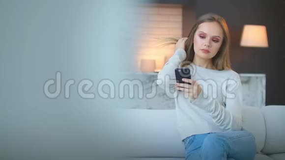 年轻漂亮的女人在公寓里坐在沙发上的智能手机上留言视频的预览图