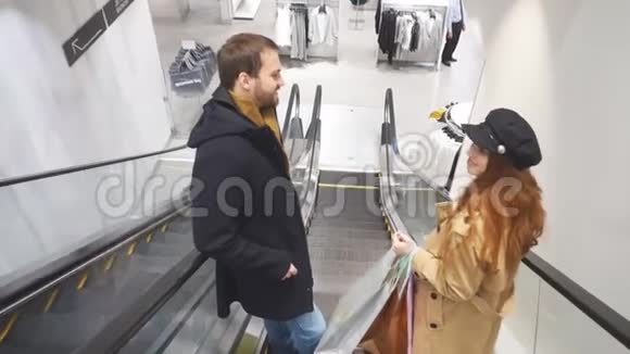 一对年轻的白种人夫妇带着购物袋站在自动扶梯上微笑视频的预览图