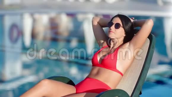 穿着红色比基尼的时尚女性在游泳池附近的日光浴甲板椅上放松视频的预览图