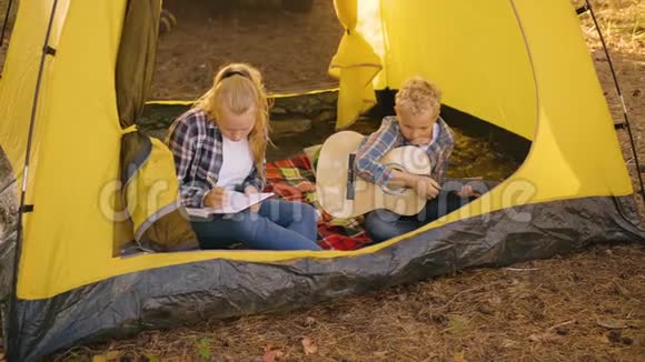 带着吉他坐在露营帐篷里的少年男孩小女孩在森林露营地的旅游帐篷里画画视频的预览图