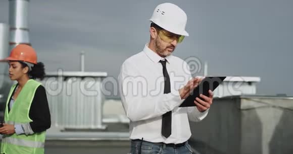 一个长得漂亮的胖乎乎的建筑工人戴着白色的头盔穿着黑色领带的衬衫他一边敲着他的平板电脑视频的预览图