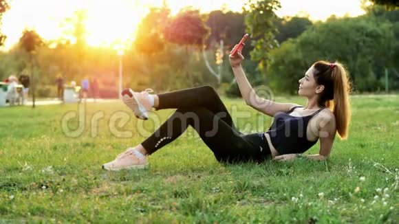 体育美女在手机上自拍日落时分躺在城市公园的草地上进行拍照视频的预览图