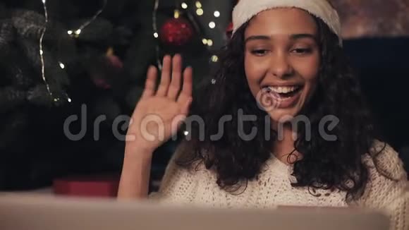 圣诞老人帽子里的年轻美女在家门口的笔记本电脑上聊天展示圣诞礼物视频的预览图