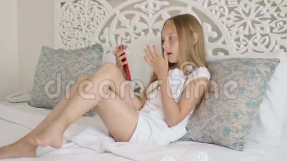 可爱的微笑女孩舔手指在卧室里使用智能手机视频的预览图