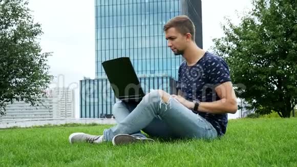 年轻的时髦男人坐在草地上用笔记本电脑在键盘上打字背景是现代的商业玻璃建筑视频的预览图