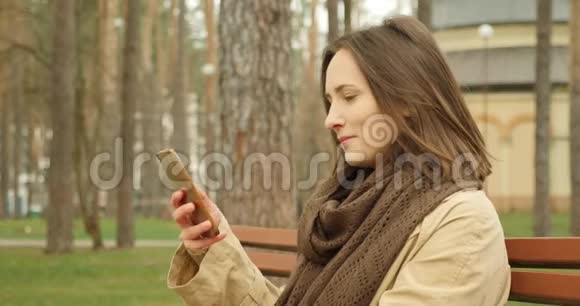 年轻的黑发女人在秋天公园坐在长凳上打电话戴着棕色围巾视频的预览图