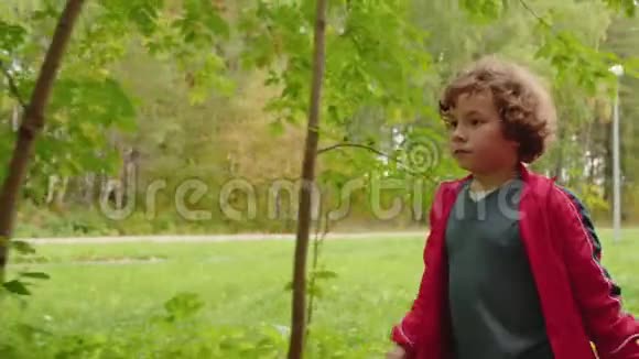 夏天公园的小卷毛男孩跑在小路上暑假在绿色公园跑步的快乐少年视频的预览图