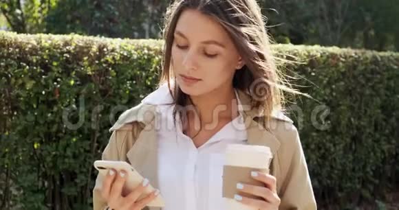 漂亮的黑发女孩呆在阳光下用智能手机拿走杯子喝热咖啡看着里面视频的预览图