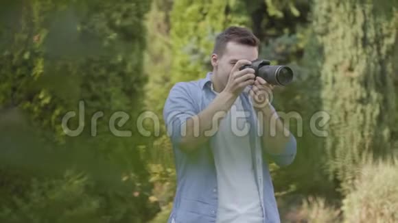 年轻的职业男性摄影师穿着休闲服装在公园或花园里用相机拍照摄影摄影视频的预览图