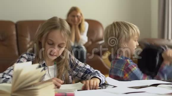 小男孩和小女孩在家学习的前景而他们的母亲坐在背景兄弟在愚弄视频的预览图