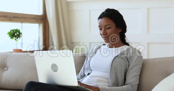 微笑的非洲女士坐在沙发上用笔记本电脑在家视频的预览图
