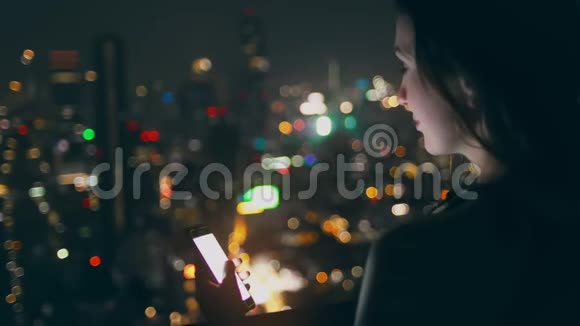 漂亮的女士手持智能手机夜城屋顶视频的预览图