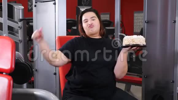 暴饮暴食的概念女肥胖女子在健身房拿着大蛋糕拇指朝下搞笑胖女孩坐健身视频的预览图