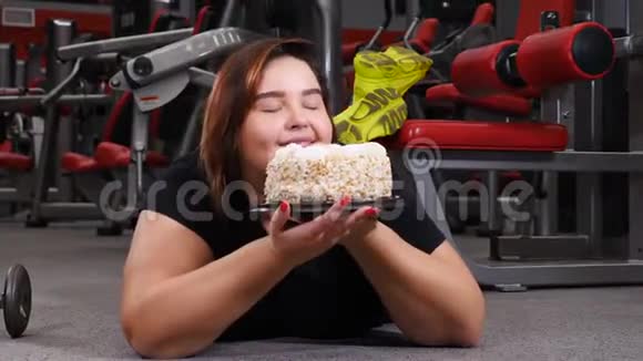 食物诱惑健身房里的肥胖女人年轻超重的女人躺在健身房的地板上拿着巨大的蛋糕意志薄弱视频的预览图