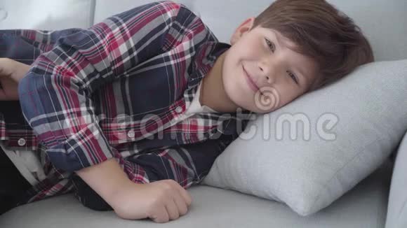 一张脸带着微笑的白种人黑发男孩的肖像灰色的眼睛躺在家里的沙发上快乐的孩子周末在室内休息视频的预览图