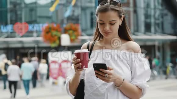在拥挤的城市街道上带着一杯咖啡的微笑可爱的女人用手机视频的预览图