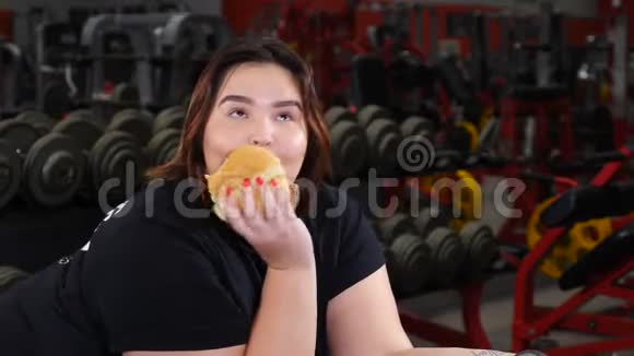 肥胖妇女的肖像手里拿着哑铃在健身房吃汉堡包微笑着看着相机食物诱惑视频的预览图