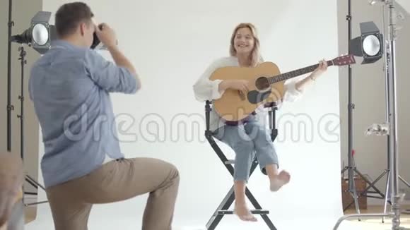 年轻的男摄影师在坐在椅子上弹吉他的白衬衫和牛仔裤的迷人女人拍照视频的预览图