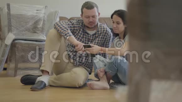 一个快乐的高加索家庭的特写镜头坐在客厅的地板上带着新的家具使用智能手机视频的预览图