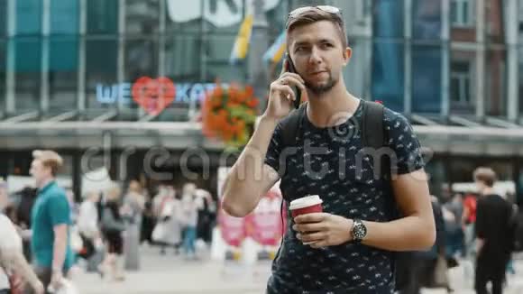 快乐的男游客带着一杯咖啡在拥挤的城市街道上用手机说话带着一个背包人们在上面视频的预览图