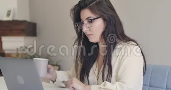 一位年轻漂亮的高加索女人戴着眼镜坐着笔记本电脑喝着茶或咖啡的特写照片视频的预览图