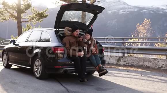 可爱的年轻的夫妇喝热茶从热水瓶坐在汽车后备箱快乐的白种人夫妇喝咖啡视频的预览图