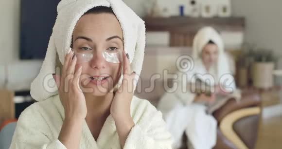 年轻可爱的高加索女人穿着白色浴袍和发巾涂着眼胶补丁穿同样衣服的模糊女孩视频的预览图