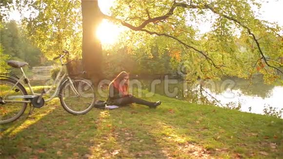 美丽的女孩在秋天公园看书放松坐在湖边的草地上骑着自行车晴天视频的预览图