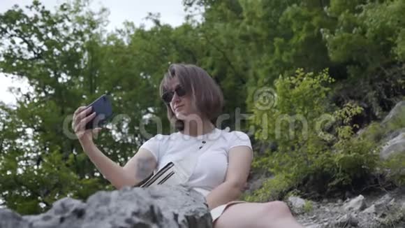 年轻的黑发女人坐在山上的岩石上在电话上自拍视频的预览图