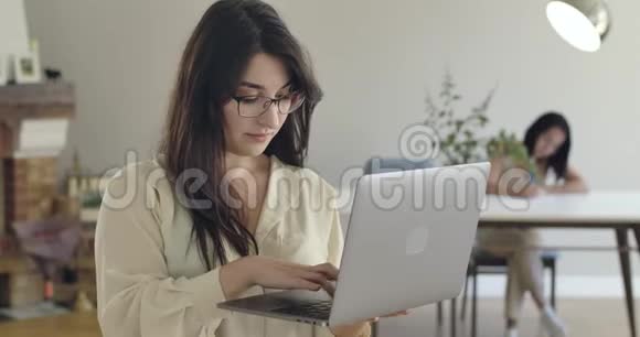 年轻漂亮的白种人女孩戴着眼镜在笔记本电脑上打字她的朋友或妹妹坐在后台视频的预览图