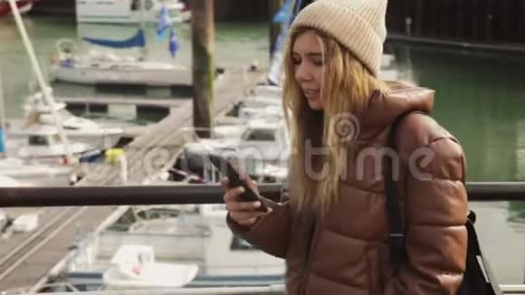 快乐的年轻迷人的时尚女孩博客作者正在港口散步看着她的手机在日落时船上视频的预览图