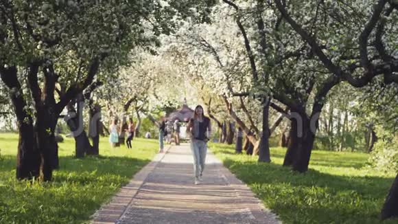 年轻的快乐女游客正独自漫步在春天阳光明媚的城市公园背景是盛开的苹果树概念视频的预览图