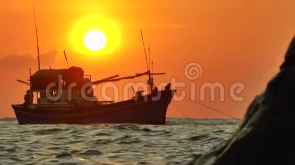 抛锚的渔船在夕阳的光线中的剪影在波浪中摆动前景是一棵岩石桦树视频的预览图