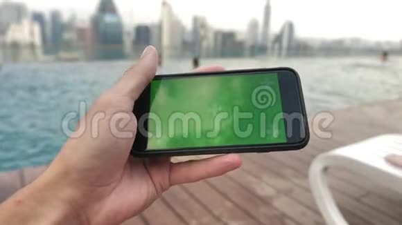 智能手机的特写镜头绿色屏幕色键在泳池附近由人以水平模式保持观看电影视频的预览图