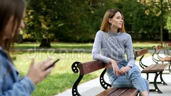 美丽的红发女人在公园的长凳上休息在模糊的前景女孩使用手机视频的预览图