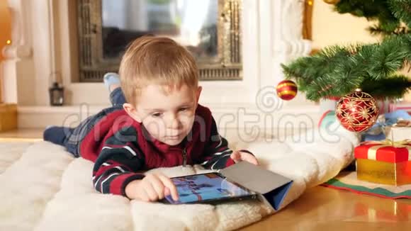 小男孩躺在圣诞树下的毯子上在数字平板电脑上玩电子游戏的4k视频儿童视频的预览图
