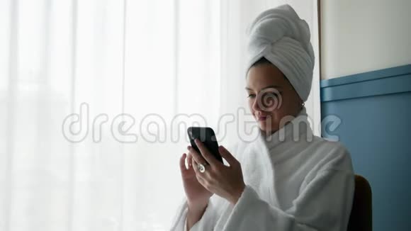 穿着浴袍头上有毛巾的女人的特写肖像使用手机视频的预览图
