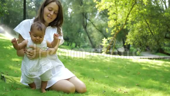 年轻漂亮的母亲教她新生的女儿在公园里的一片绿色的新鲜草地上行走年轻的母亲快乐视频的预览图