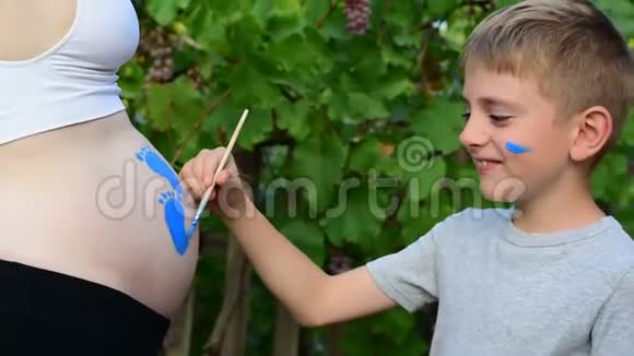 微笑的小男孩用刷子在他怀孕妈妈宝宝的肚子上画脚印怀孕的概念关门视频的预览图