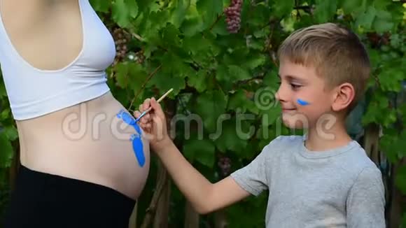 笑的男孩用刷子在他怀孕妈妈宝宝的肚子上画脚印怀孕的概念特写视频的预览图