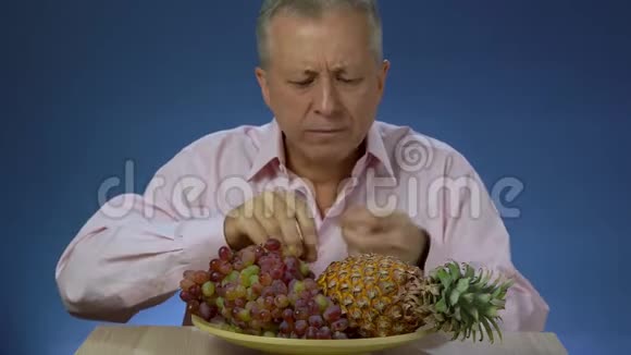 一个穿着衬衫的中年男人迅速而快乐地吃着葡萄葡萄躺在一个大菠萝旁边的一个大盘子上视频的预览图