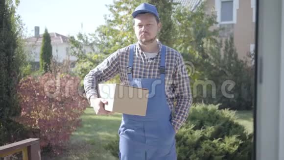 一位年轻的白人男性快递员穿着蓝色工作服给女性顾客送盒子并要求付款的肖像视频的预览图