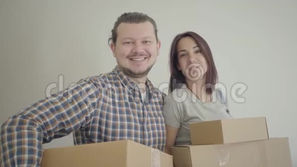 微笑白人夫妇站在纸板箱和看相机幸福的年轻家庭搬到他们的新家视频的预览图